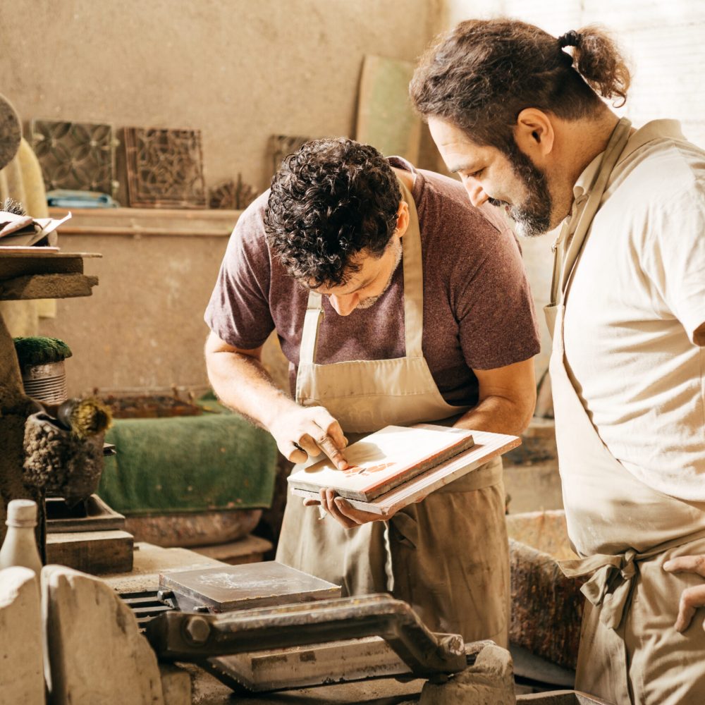 Two factory workers meticulously examine a mosaic cement tile at a manufacturing facility, ensuring its quality and precision. They are focused and attentive, collaborating on the inspection.