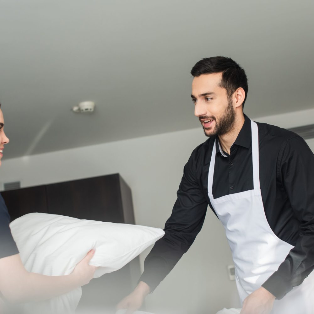 young positive housekeepers changing bedding in modern hotel room